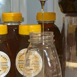 Puede incluir: Un tarro de vidrio transparente se llena de miel de un dispensador. Varias botellas de miel de color &aacute;mbar con tapas amarillas est&aacute;n en el fondo. Las etiquetas dicen "PIARY HONEY" y "LEASBURG PURE HONEY, Peso neto 227g."