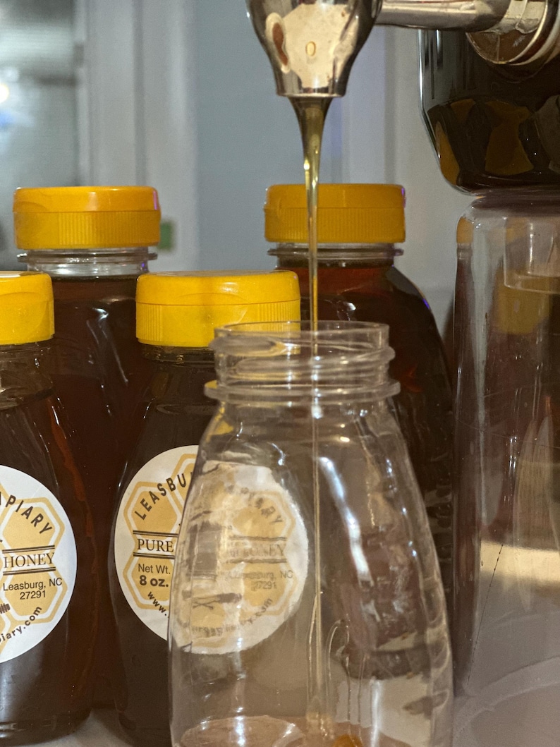 Puede incluir: Un tarro de vidrio transparente se llena de miel de un dispensador. Varias botellas de miel de color &aacute;mbar con tapas amarillas est&aacute;n en el fondo. Las etiquetas dicen "PIARY HONEY" y "LEASBURG PURE HONEY, Peso neto 227g."