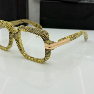 Puede incluir: Gafas con un diseño único de piel de serpiente en tonos amarillos, marrones y negros. Las monturas tienen forma cuadrada con un detalle metálico dorado en las patillas. Las lentes son transparentes.