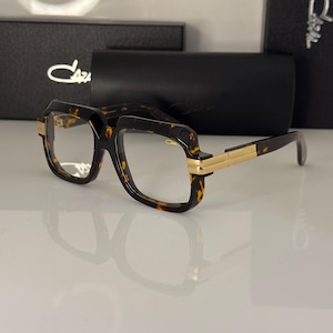 May include: Tortoiseshell-patterned spectacles with a thick, square-shaped frame and clear lenses. The temples feature gold-tone metal accents, and the brand name Cazal is printed on the upper right lens. A black protective case is positioned behind.