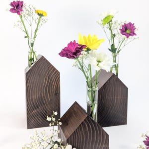 May include: Dark brown wooden house-shaped vases, each holding a small glass tube with colourful flowers. The flowers include purple, yellow, and white daisies. The vases are arranged against a white backdrop.