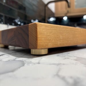 Könnte beinhalten: Ein rechteckiges Holzschneidebrett mit zweifarbigem Design. Eine Seite ist dunkelbraun, die andere hellbraun. Das Brett ruht auf vier kleinen, goldfarbenen Füßen. Das Brett liegt auf einer weißen Marmorarbeitsplatte.