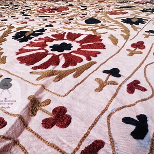 Op de afbeelding: Een lichtroze textiel met ingewikkelde, met de hand geborduurde bloemmotieven in donkerrood, zwart en goudbruin garen. De stof toont een herhalend patroon van grote centrale bloesems omringd door kleinere takjes en golvende lijnen.