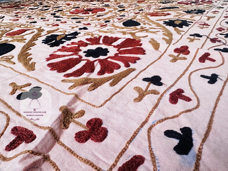 Op de afbeelding: Een lichtroze textiel met ingewikkelde, met de hand geborduurde bloemmotieven in donkerrood, zwart en goudbruin garen. De stof toont een herhalend patroon van grote centrale bloesems omringd door kleinere takjes en golvende lijnen.
