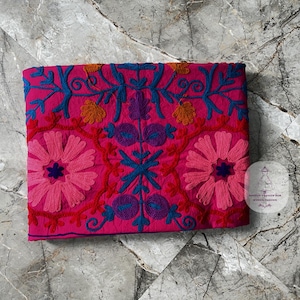 May include: A vibrant pink textile with intricate embroidery. The design features large pink flowers, blue vines, and accents of orange and purple. The textile is folded and lies on a marble surface.
