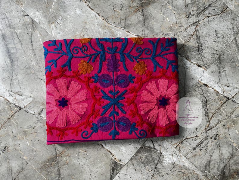 May include: A vibrant pink textile with intricate embroidery. The design features large pink flowers, blue vines, and accents of orange and purple. The textile is folded and lies on a marble surface.