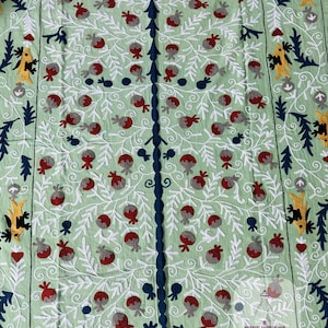 Puede incluir: Textil verde claro con un diseño bordado de árbol de la vida. El árbol presenta un tronco y ramas azul oscuro, con frutos rojos y grises y hojas blancas. El borde tiene elementos decorativos amarillos y azules. El textil tiene el texto "MANSURI FASHION HUB WOMAN FASHION".