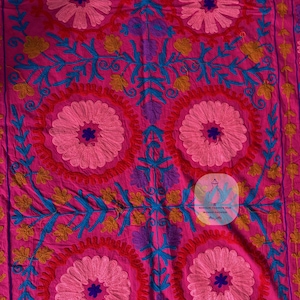 May include: A vibrant pink textile with intricate embroidery. The design features large, circular floral patterns in shades of pink, blue, yellow, and purple. The embroidery is detailed and creates a textured surface, showcasing a traditional craft.