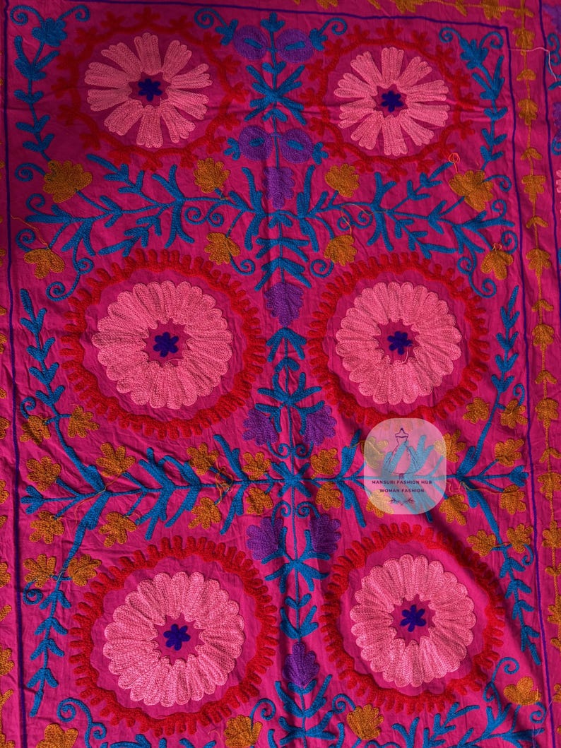 May include: A vibrant pink textile with intricate embroidery. The design features large, circular floral patterns in shades of pink, blue, yellow, and purple. The embroidery is detailed and creates a textured surface, showcasing a traditional craft.