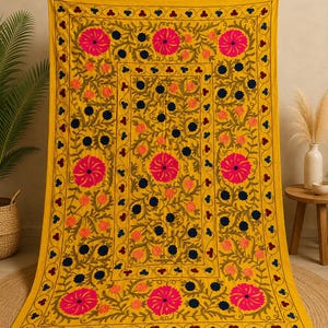 May include: A rectangular yellow textile featuring intricate embroidery with large hot pink circular floral motifs, smaller dark blue flowers, and orange accents. The design includes scrolling green vines and a decorative border with fringe.