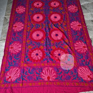 May include: A large, rectangular textile in shades of pink, fuchsia, and purple, featuring intricate floral embroidery. The design includes large, stylized flowers and detailed leaf patterns, creating a vibrant and ornate appearance.