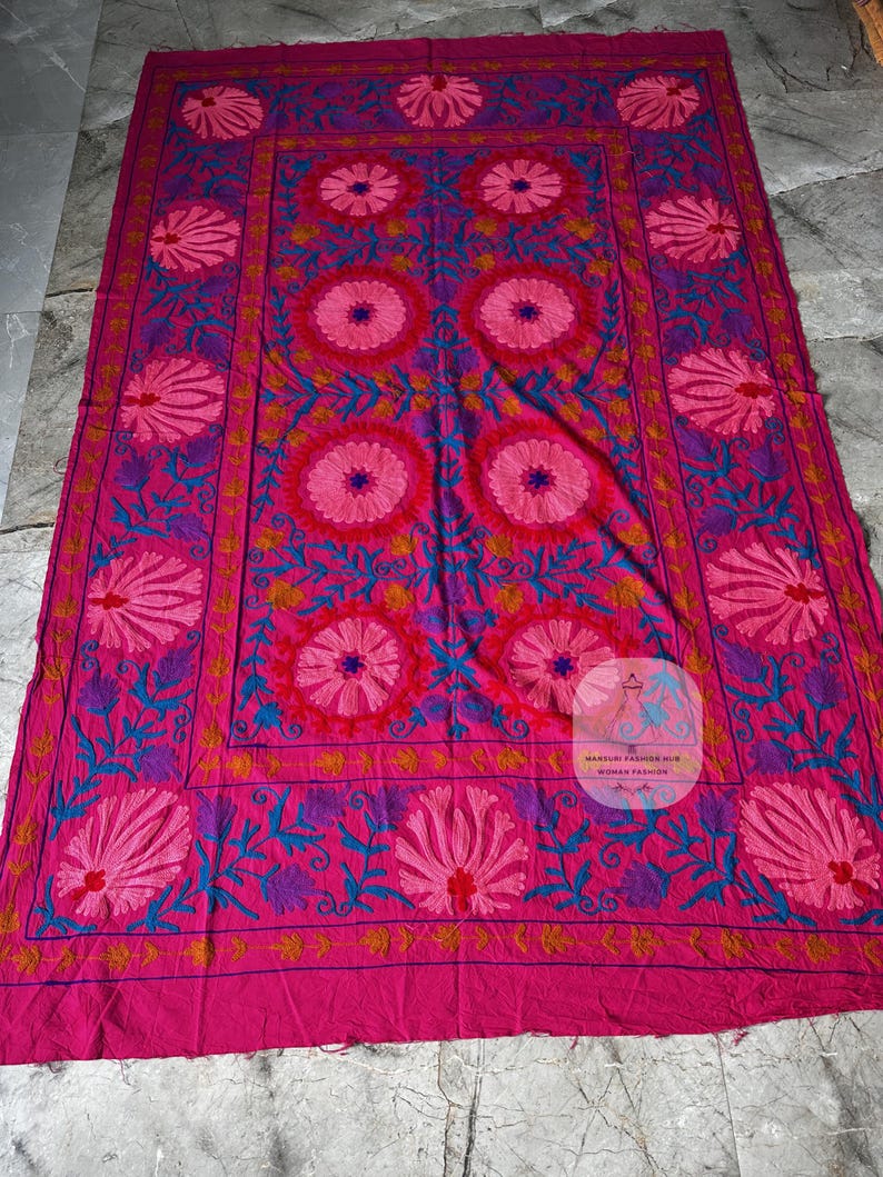 May include: A large, rectangular textile in shades of pink, fuchsia, and purple, featuring intricate floral embroidery. The design includes large, stylized flowers and detailed leaf patterns, creating a vibrant and ornate appearance.