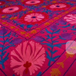 May include: A vibrant pink textile with intricate embroidery. The design features large floral patterns in shades of pink, red, and white, accented with teal, purple, and yellow details. The textile is likely a decorative piece, such as a bedspread or wall hanging.
