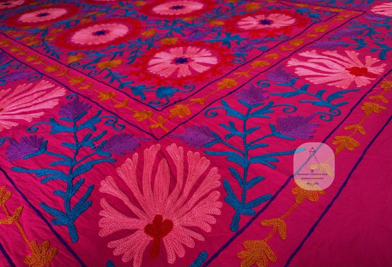 May include: A vibrant pink textile with intricate embroidery. The design features large floral patterns in shades of pink, red, and white, accented with teal, purple, and yellow details. The textile is likely a decorative piece, such as a bedspread or wall hanging.