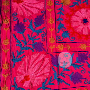 May include: Bright pink textile with intricate floral embroidery in shades of pink, purple, blue, and gold. The design features large stylized flowers and scrolling vines, creating a vibrant and detailed pattern. The textile has a label that says "MANDURI FASHION HUB WOMAN FASHION".