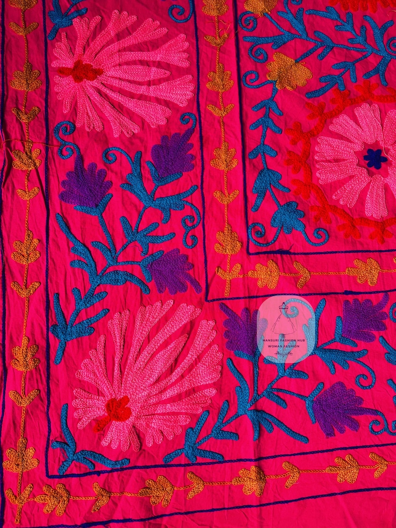 May include: Bright pink textile with intricate floral embroidery in shades of pink, purple, blue, and gold. The design features large stylized flowers and scrolling vines, creating a vibrant and detailed pattern. The textile has a label that says "MANDURI FASHION HUB WOMAN FASHION".