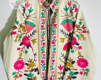 Könnte beinhalten: Eine cremeweiße Jacke mit leuchtend floralen Stickereien in Pink, Türkis und Senfgelb. Das Kleidungsstück verfügt über einen Stehkragen, lange Ärmel und eine Vorderöffnung, die ein schwarz-weiß gestreiftes Innenfutter mit kleinen roten Herzen zeigt.