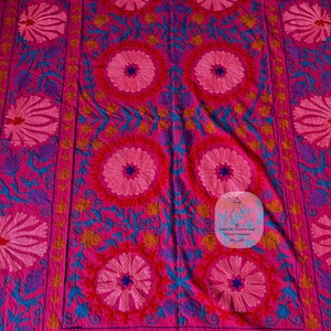 May include: A vibrant pink textile featuring intricate floral embroidery in shades of pink, blue, purple, and yellow. The design includes large central circular flower patterns and detailed border elements, creating a rich, decorative appearance.
