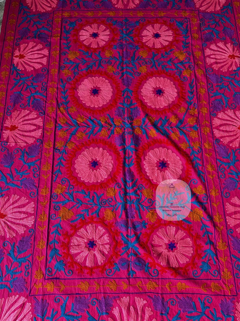 May include: A vibrant pink textile featuring intricate floral embroidery in shades of pink, blue, purple, and yellow. The design includes large central circular flower patterns and detailed border elements, creating a rich, decorative appearance.