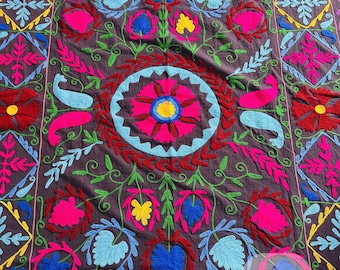 May include: A detailed textile with a dark brown background, featuring vibrant embroidery in shades of pink, red, blue, yellow, and green. The design includes floral and geometric patterns, creating a rich and colorful tapestry.