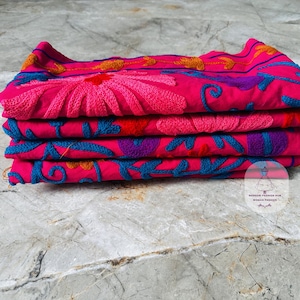 May include: A stack of four folded textiles in vibrant pink, adorned with intricate floral embroidery. The embroidery features shades of pink, blue, purple, and orange, creating a colorful and detailed design.