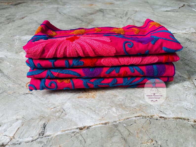 May include: A stack of four folded textiles in vibrant pink, adorned with intricate floral embroidery. The embroidery features shades of pink, blue, purple, and orange, creating a colorful and detailed design.