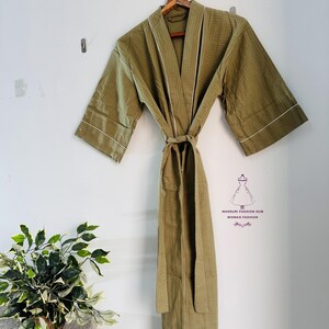 May include: Olive green waffle-weave robe with white trim, hanging on a wooden hanger. The robe has a matching belt and is displayed against a white wall. The robe is a comfortable and stylish garment.