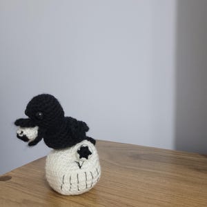 Könnte beinhalten: Eine handgefertigte Häkelpuppe mit einer schwarzen Kreatur, die auf einem weißen Schädel sitzt. Der Schädel hat schwarze gestickte Details für Augen, Nase und Mund. Die Puppe befindet sich auf einer Holzoberfläche.