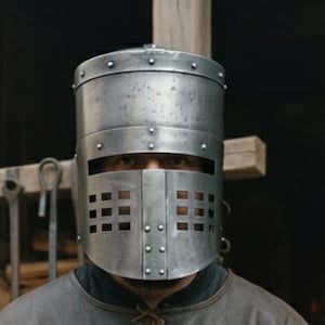 Op de afbeelding: Een persoon draagt een helm in middeleeuwse stijl en leren harnas. De helm is zilverkleurig met een rechthoekige gezichtsopening. Het harnas is bruin met een riem. De achtergrond toont een smederij met gereedschap en vuur.