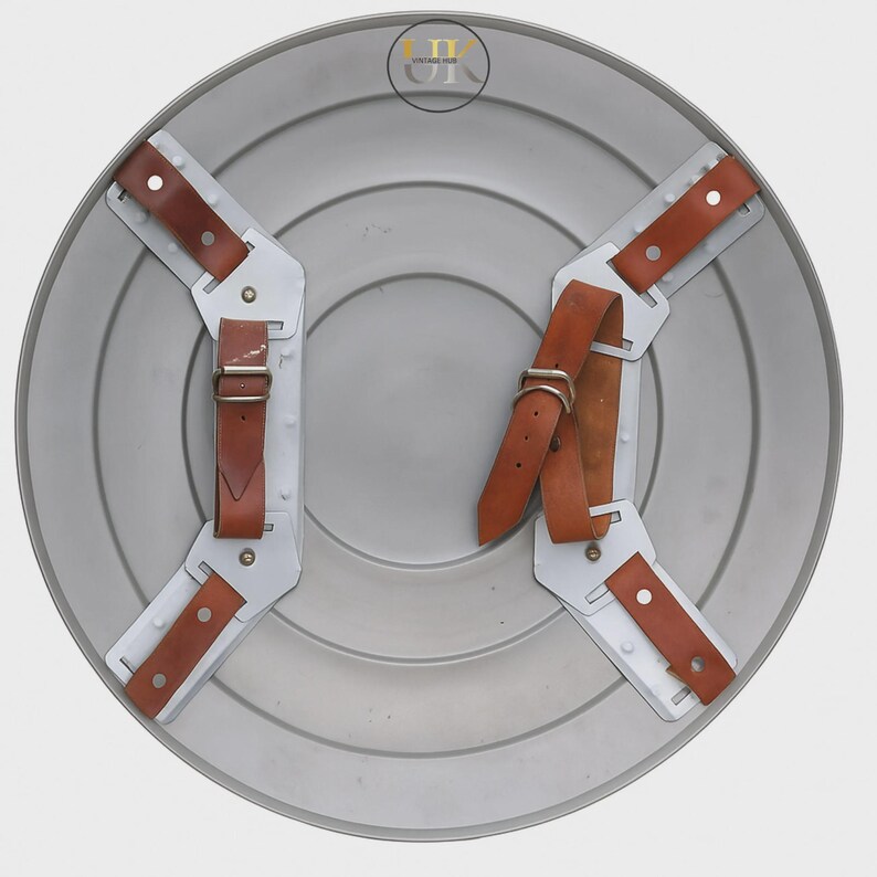 May include: A silver-coloured, circular shield with concentric rings. It features four white metal brackets with brown leather straps and buckles for securing the shield. The shield has a vintage appearance.