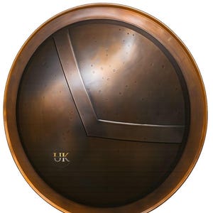 May include: A round, antique-style shield with a bronze finish. The shield features a dark center with a lighter bronze border and a diagonal metal accent. The letters "UK" are visible near the bottom. The shield has a slightly weathered appearance.
