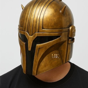 Puede incluir: Casco dorado con visera negra y crestas verticales. El casco tiene un acabado met&aacute;lico y un logotipo en la parte inferior derecha. La persona lleva una camiseta negra.