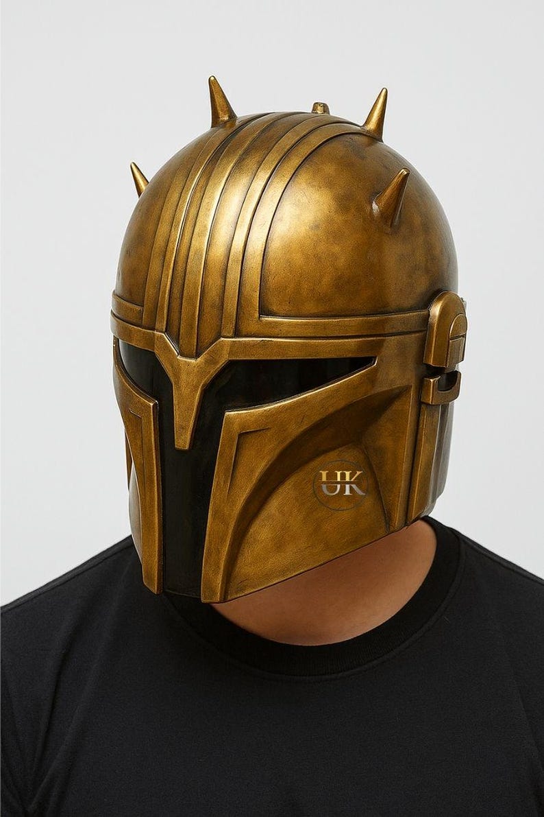 Puede incluir: Casco dorado con visera negra y crestas verticales. El casco tiene un acabado met&aacute;lico y un logotipo en la parte inferior derecha. La persona lleva una camiseta negra.
