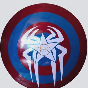 May include: A circular shield in red, white, and blue. The design features a white star at the centre, with a spider-like pattern and the text "VINTAGE HUB" in a circle. This shield is likely a decorative item or a collectible.