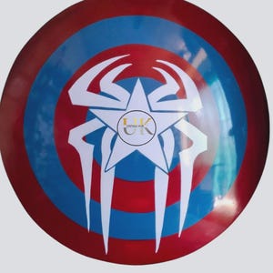 May include: A circular shield with a red, white, and blue design. The shield features a central white star with a spider-like design and the text "UK VINTAGE HUB". The shield has a glossy finish.