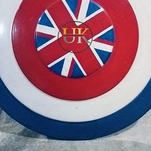 May include: A round shield with a blue, white, and red color scheme. The shield features a central red circle with a white and blue starburst design. The outer ring is white, and the edge is blue.