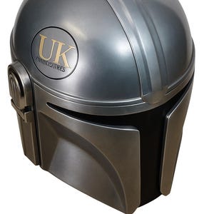 May include: A metallic silver Mandalorian helmet with a black visor. The helmet has a gold emblem with the letters "UK" and the word "OVNICISATRES". The helmet has a smooth, rounded design with a vertical ridge.