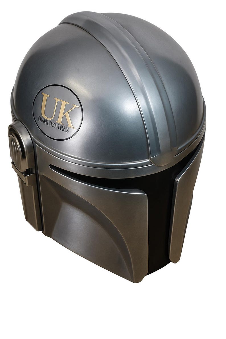 May include: A metallic silver Mandalorian helmet with a black visor. The helmet has a gold emblem with the letters "UK" and the word "OVNICISATRES". The helmet has a smooth, rounded design with a vertical ridge.