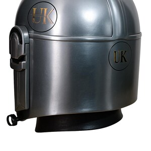May include: A metallic gray helmet with a rounded top and a black base. The helmet features a visor and the letters "UK" in a gold color on both sides. The design is reminiscent of a futuristic or sci-fi aesthetic.