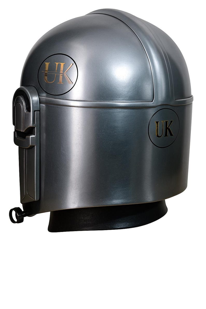 May include: A metallic gray helmet with a rounded top and a black base. The helmet features a visor and the letters "UK" in a gold color on both sides. The design is reminiscent of a futuristic or sci-fi aesthetic.