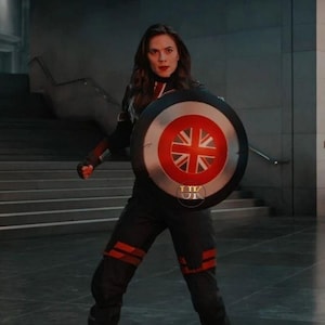 May include: A person in a black and red uniform holding a shield with a Union Jack design. The shield has a red and white border. The person is standing in a modern building with a staircase.