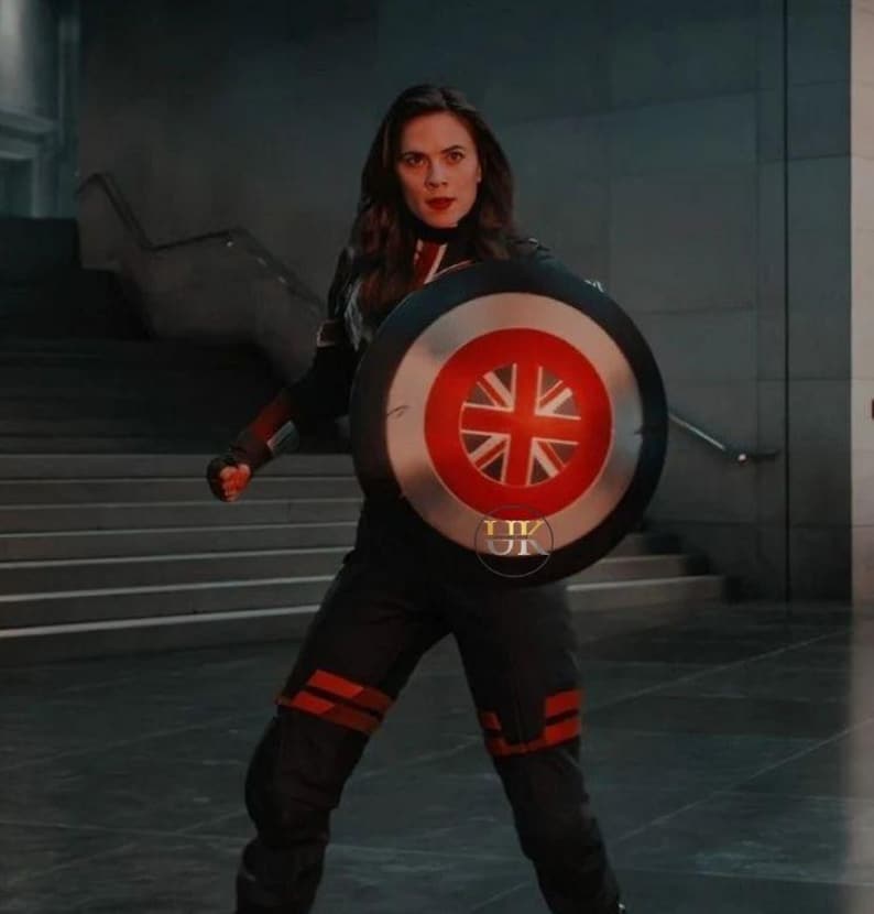 May include: A person in a black and red uniform holding a shield with a Union Jack design. The shield has a red and white border. The person is standing in a modern building with a staircase.