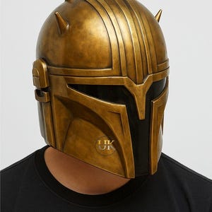 Puede incluir: Un casco dorado con un dise&ntilde;o distintivo, con crestas verticales y cuatro protuberancias puntiagudas. El casco tiene una visera oscura y un acabado met&aacute;lico, lo que sugiere un tema futurista o de fantas&iacute;a.