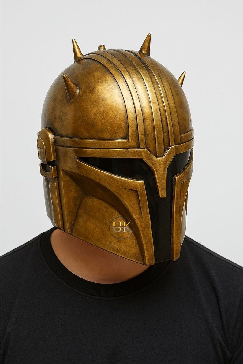 Puede incluir: Un casco dorado con un dise&ntilde;o distintivo, con crestas verticales y cuatro protuberancias puntiagudas. El casco tiene una visera oscura y un acabado met&aacute;lico, lo que sugiere un tema futurista o de fantas&iacute;a.