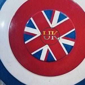 May include: A round shield with a patriotic design. The shield features concentric circles in blue, white, and red. The center showcases a red circle with a Union Jack design, incorporating blue, white, and red elements. The shield has a glossy finish.
