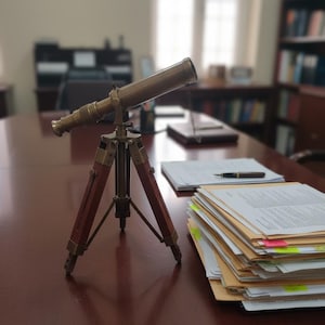 Könnte beinhalten: Ein Messing-Teleskop auf einem Holzstativ, das auf einem dunkelbraunen Tisch steht. Das Teleskop ist nach oben geneigt, mit einem Stapel Papiere und einem Stift in der Nähe. Der Hintergrund ist unscharf, was auf eine Büroumgebung hindeutet.
