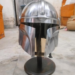 May include: A silver-colored helmet with a visor, displayed on a black stand. The helmet has a rounded top with ridges and a wide, angular visor. The stand is circular and made of dark wood.