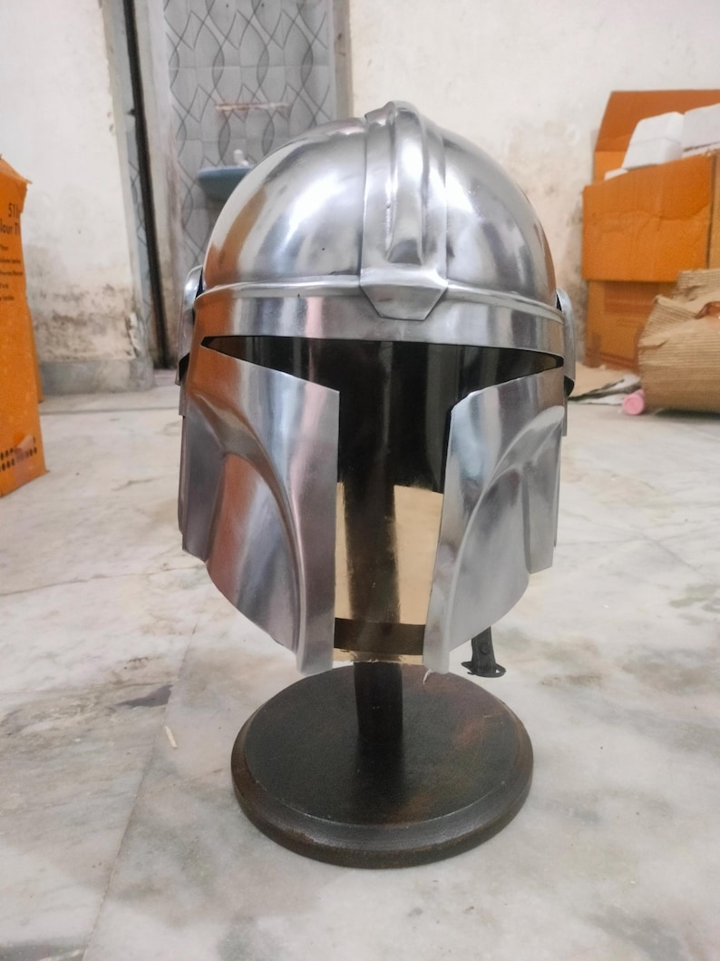 May include: A silver-colored helmet with a visor, displayed on a black stand. The helmet has a rounded top with ridges and a wide, angular visor. The stand is circular and made of dark wood.