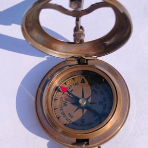 May include: A vintage brass compass with a hinged lid, featuring a directional dial with cardinal points and degree markings. The compass has a red needle and a loop for hanging. The image shows the compass open, casting a shadow.