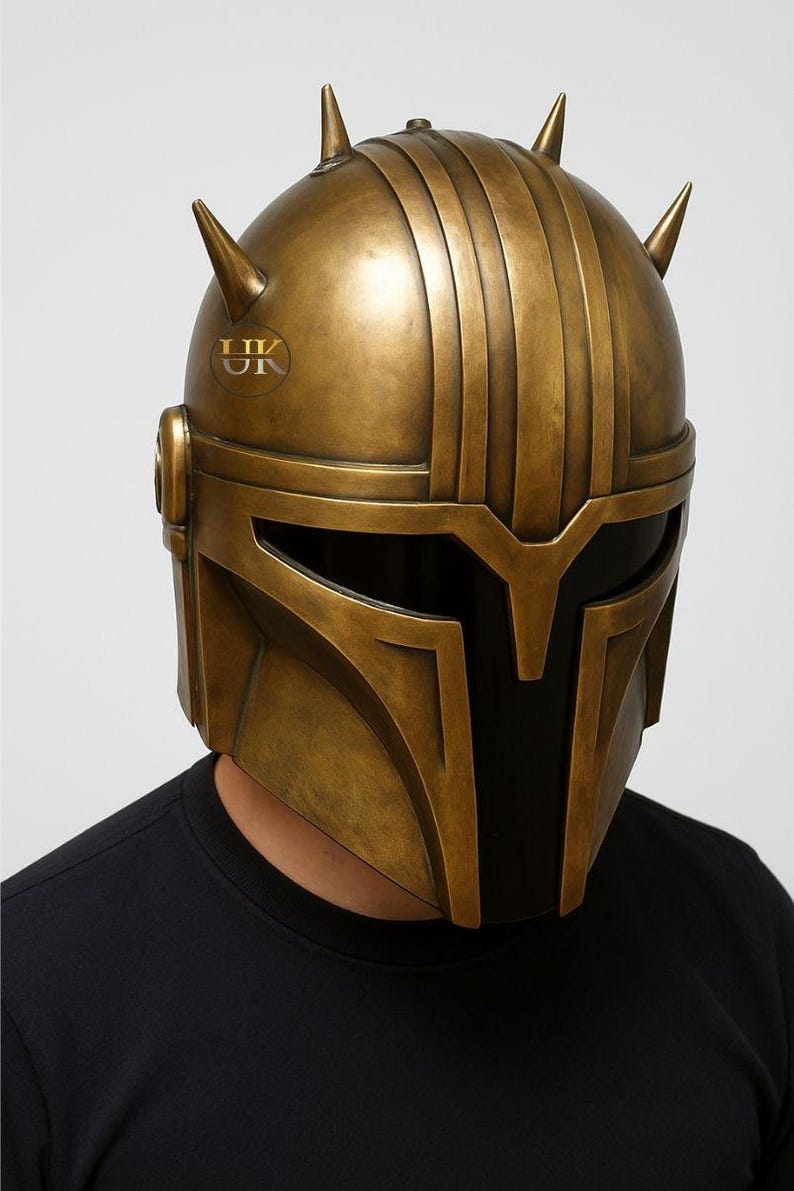 Puede incluir: Un casco de color bronce con un dise&ntilde;o distintivo, con crestas verticales y p&uacute;as puntiagudas. El casco tiene una visera y un logotipo que dice "UK". El casco lo lleva una persona que lleva una camiseta negra.